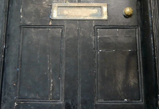 A wooden black door with faded and peeling paint, with rusting knob and mail slot